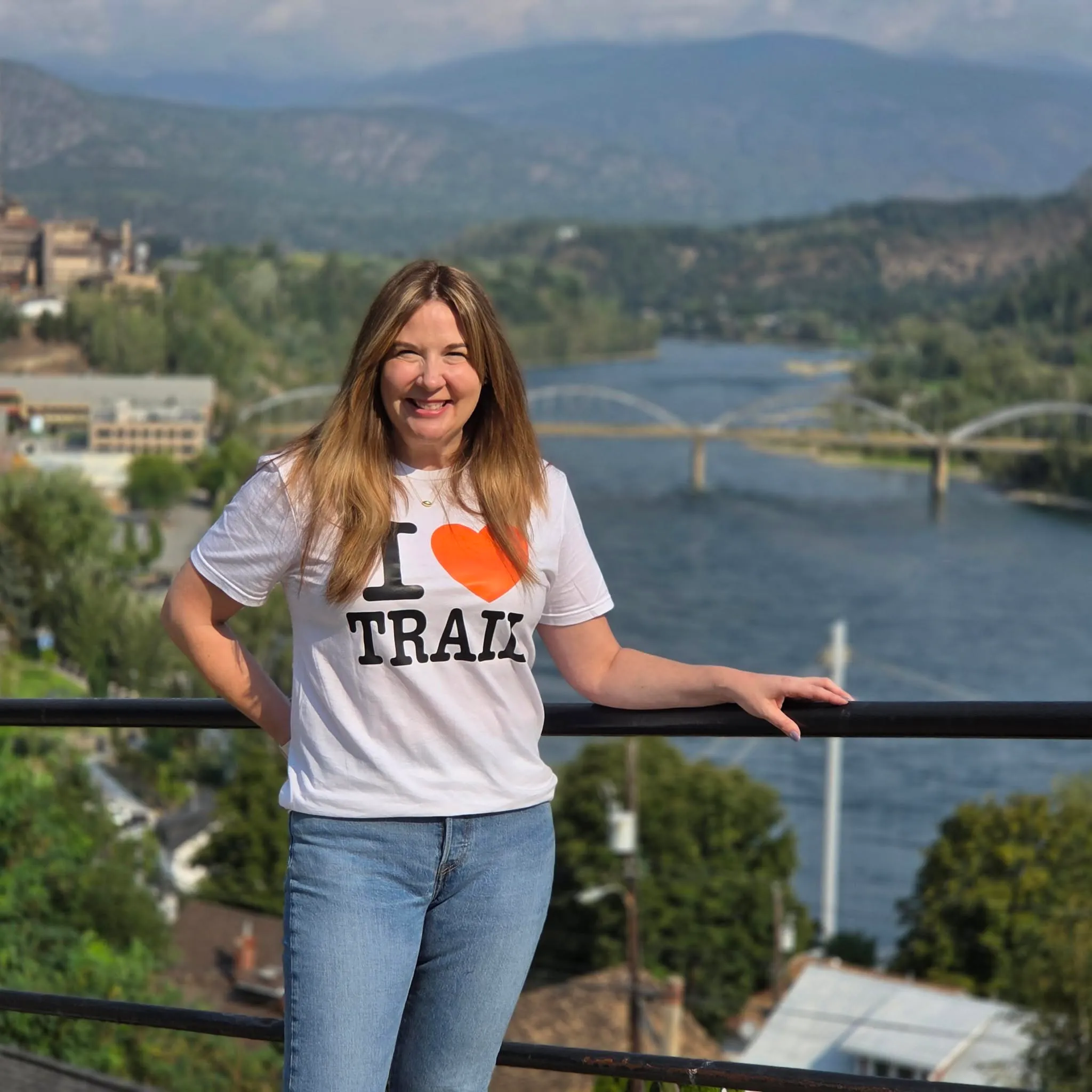 Bev Benson smiling in an I ♥ Trail t-shirt, with the Columbia River and Trail's bridges behind her